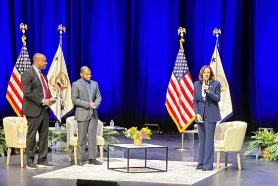 photo of three people on stage with flags, chairs, table